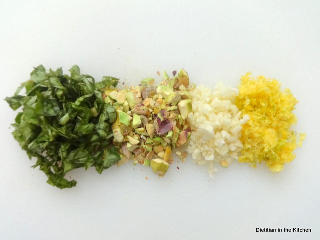 A cutting board with piles of chopped basil, pistachios, garlic, and lemon zest for making gremolata.