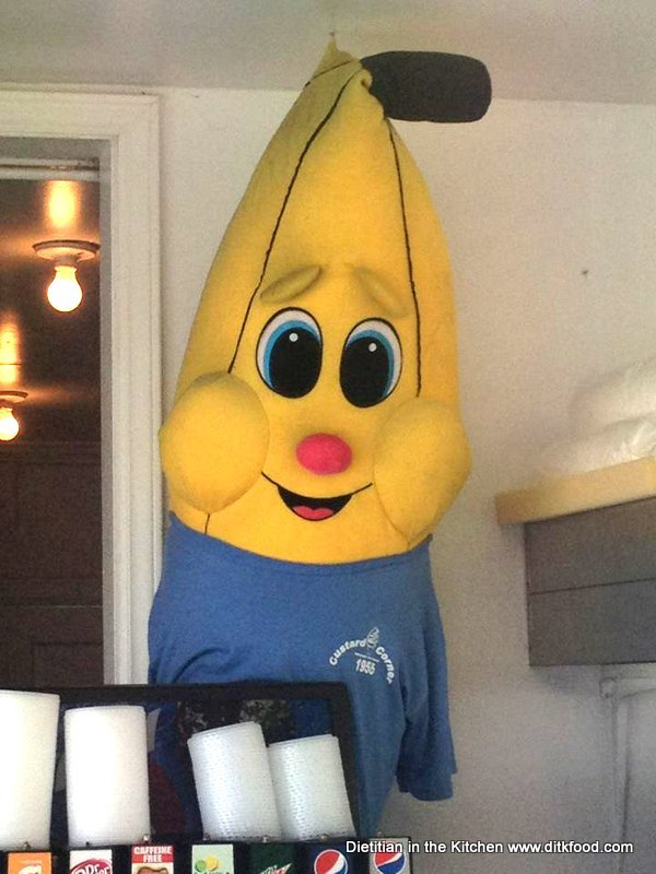 A giant stuffed banana with a blue tee shirt hanging in an ice cream shop by a drink machine. 