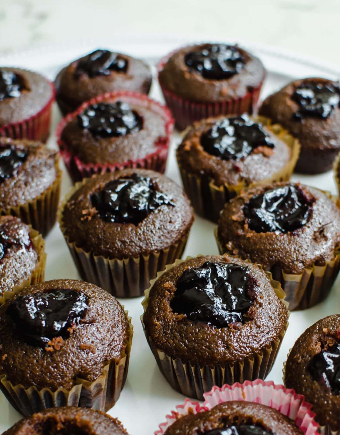 A close up shot of chocolate cupcakes on a white platter with the center of the cupcakes cut out and filled with cherry filling. 