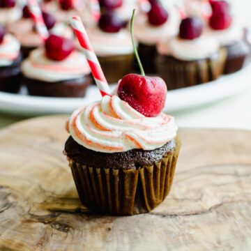 A close-up shot of a chocolate cupcake topped with red striped frosting, a cherry, and a red striped paper straw.