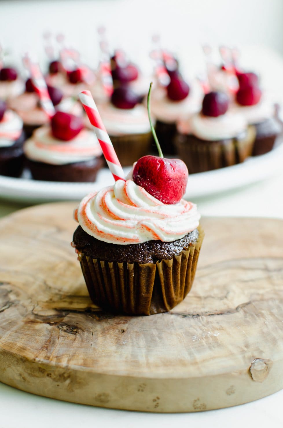 A close-up shot of a chocolate cupcake topped with red striped frosting, a cherry, and a red striped paper straw. 