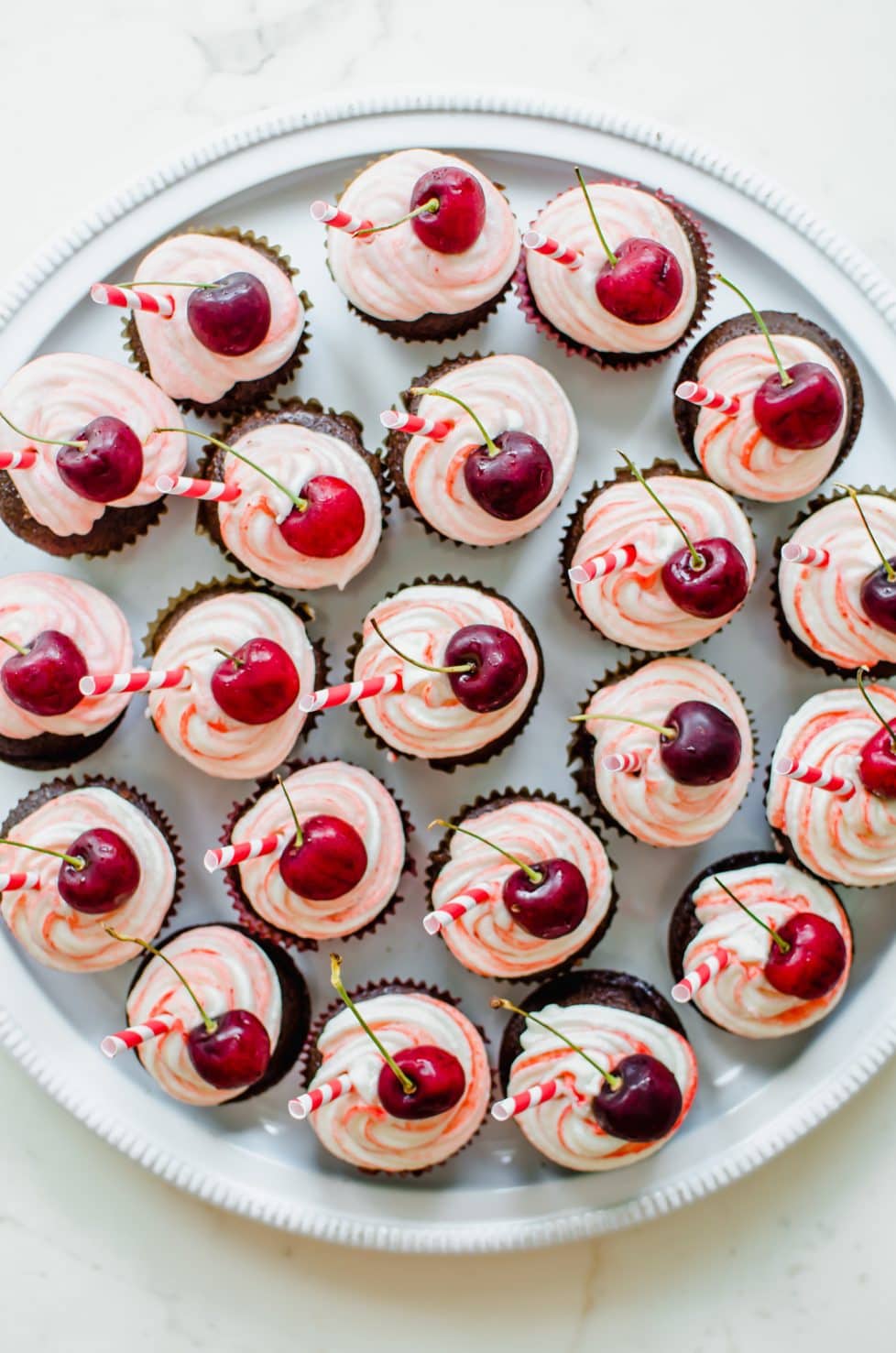 An overhead shot of a large platter of chocolate cupcakes topped with red striped frosting, a cherry, and a red striped paper straw. 