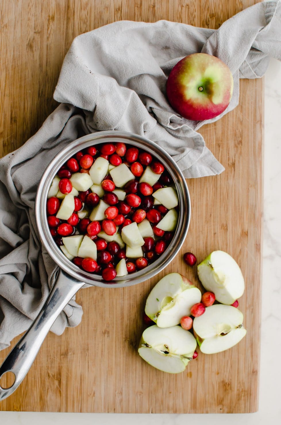 A saucepan with fresh cranberries and sliced apple sitting on a cutting board with a grey dish towel underneath.