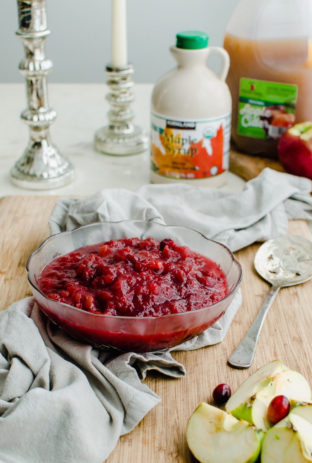 A glass bowl of apple cranberry sauce sitting on a cutting board with a spoon and sliced apples on the side. 