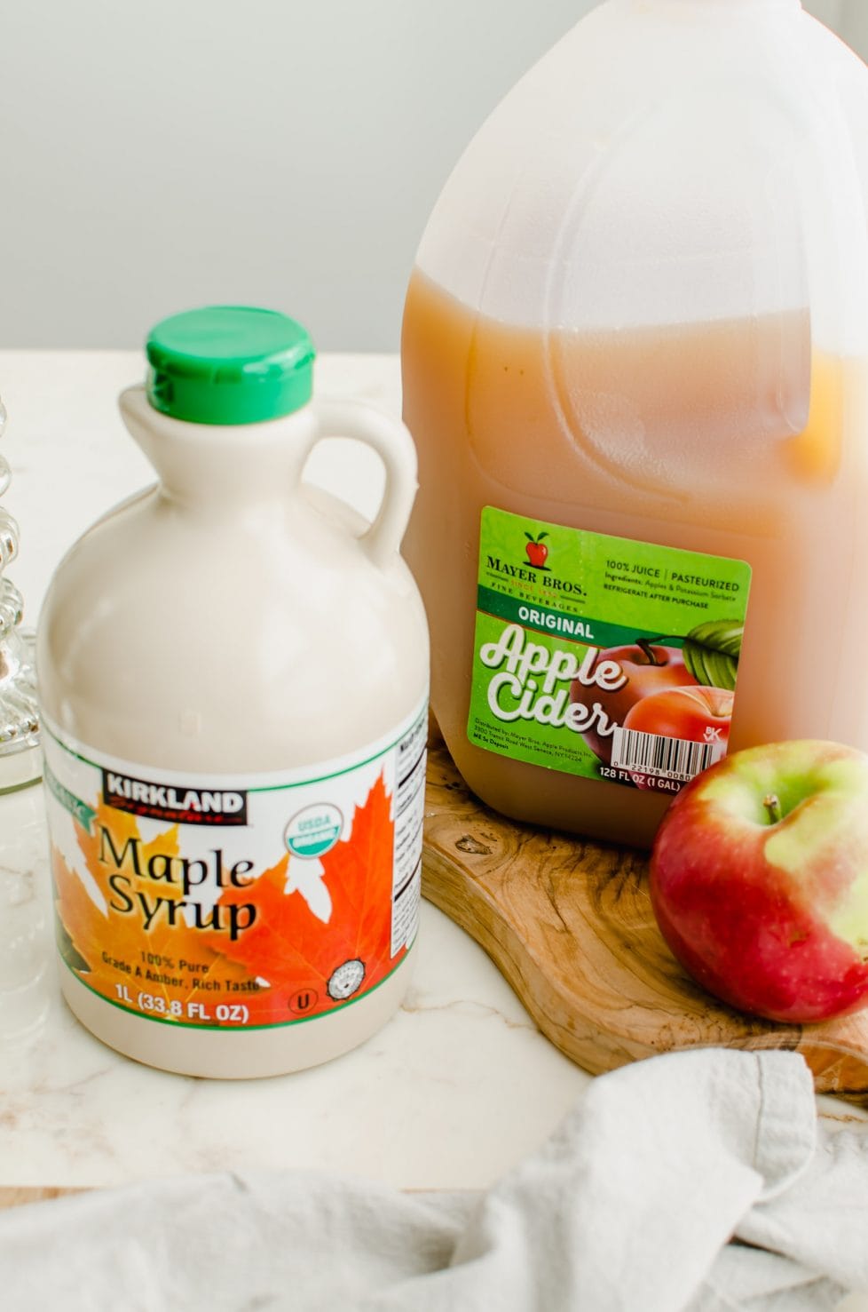 A jug of apple cider, bottle of maple syrup, and an apple sitting on a cutting board.