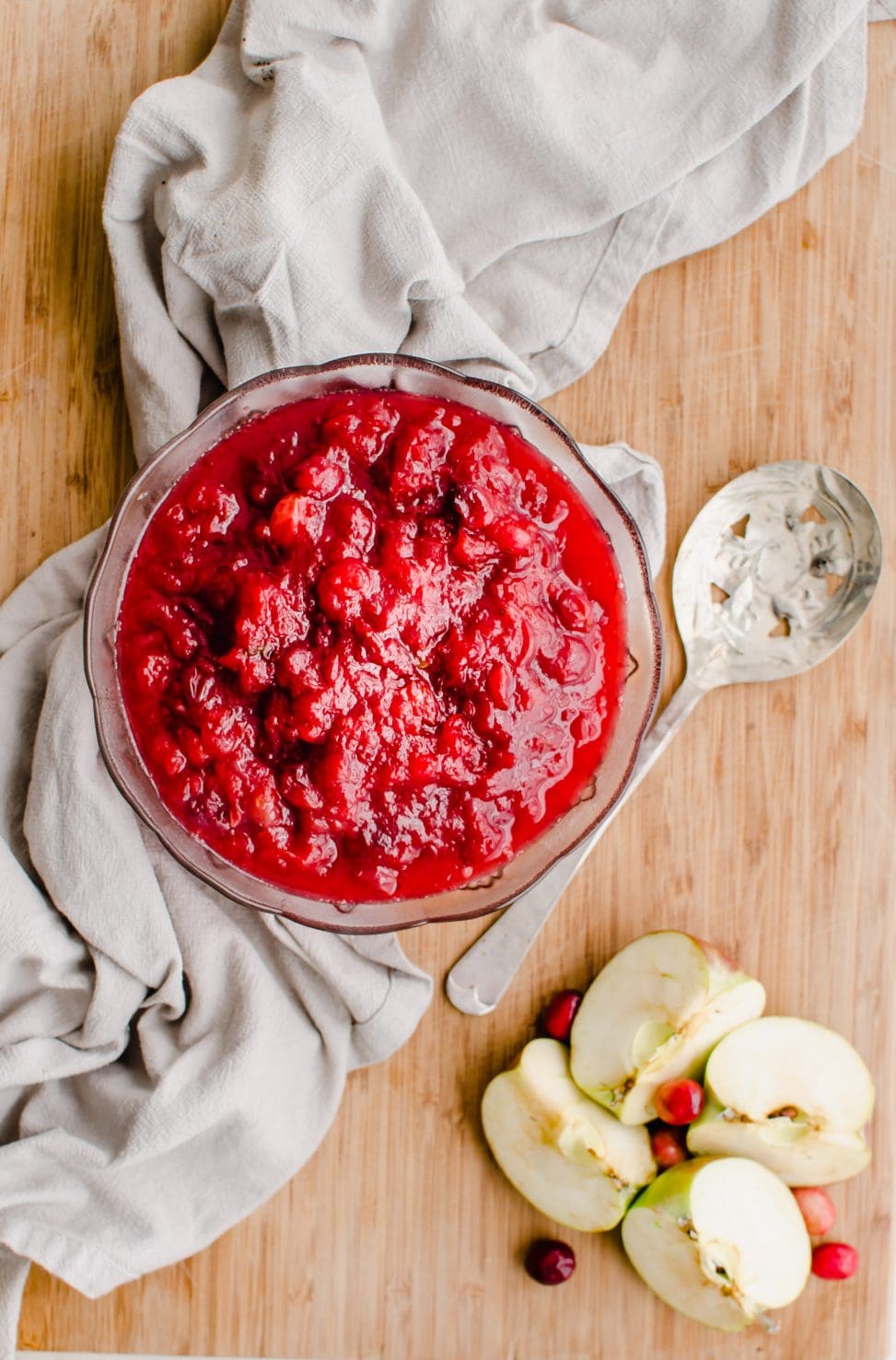 A glass bowl of apple cranberry sauce sitting on a cutting board with a spoon and sliced apples on the side. 