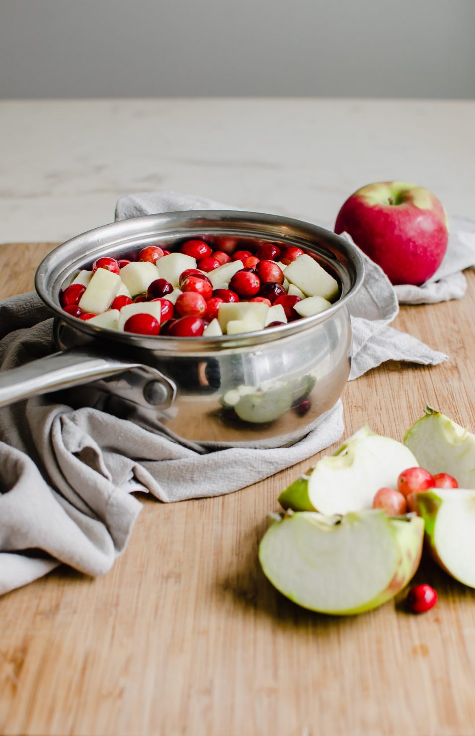 A saucepan with fresh cranberries and sliced apple sitting on a cutting board with a grey dish towel underneath.