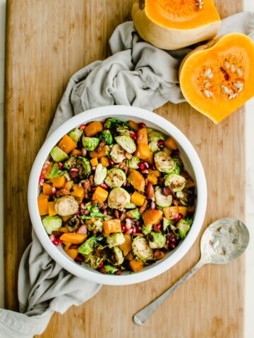 A white bowl filled with roasted brussels sprouts and butternut squash with a cut butternut squash half on the side.