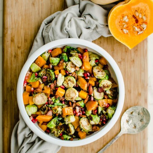 A white bowl filled with roasted brussels sprouts and butternut squash with a cut butternut squash half on the side.