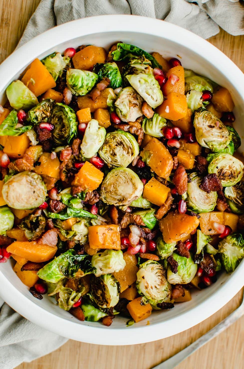 A close-up overhead shot of a white bowl filled with roasted brussels sprouts, butternut squash, and pomegranate arils.