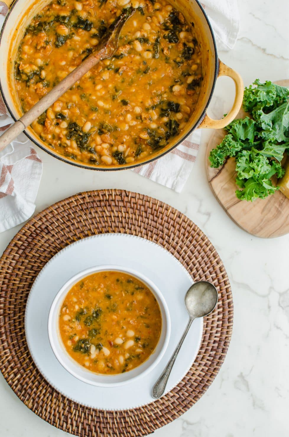 A Dutch oven and white soup bowl filled with 5-Ingredient Italian Bean Stew with a bunch of kale on the side.
