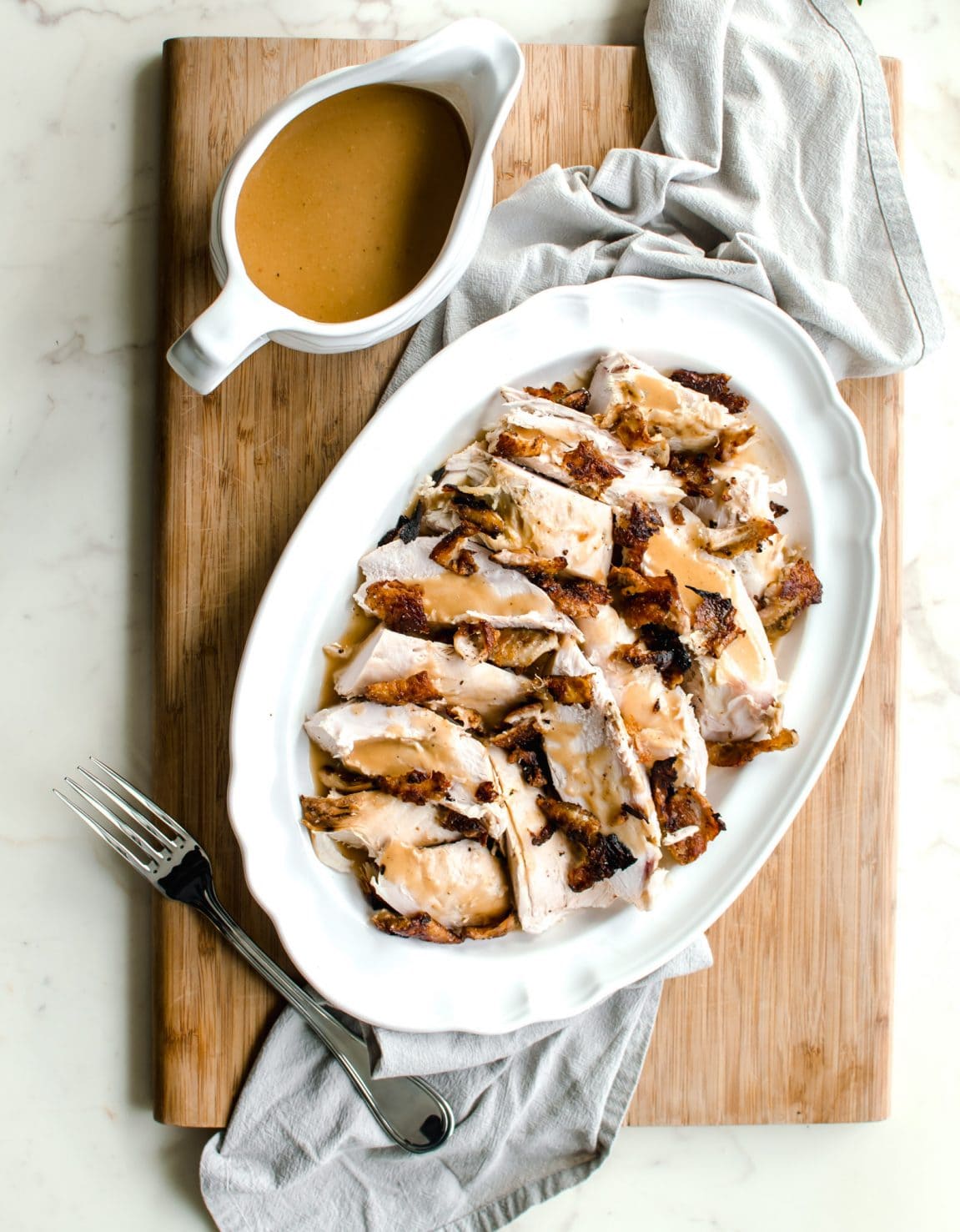 A white platter filled with turkey breast that is topped with crispy skin and gravy with a dish of gravy on the side.