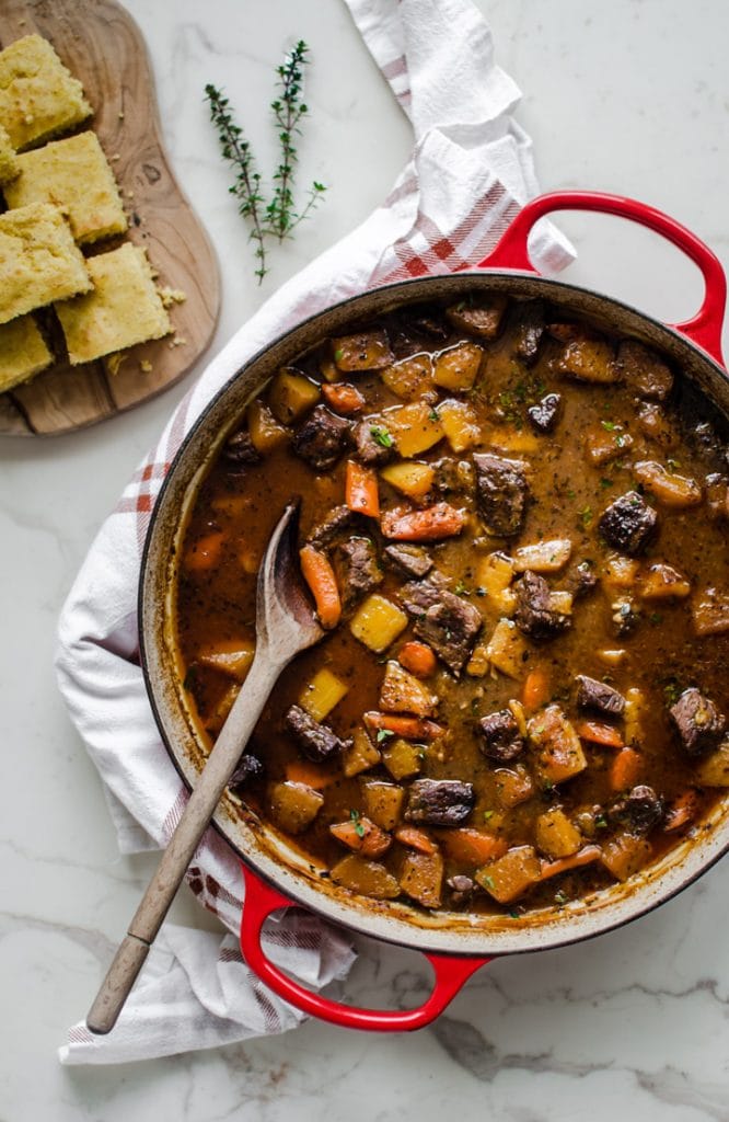 A red cast iron braised filled with beef stew and a cutting board with cornbread on the side.