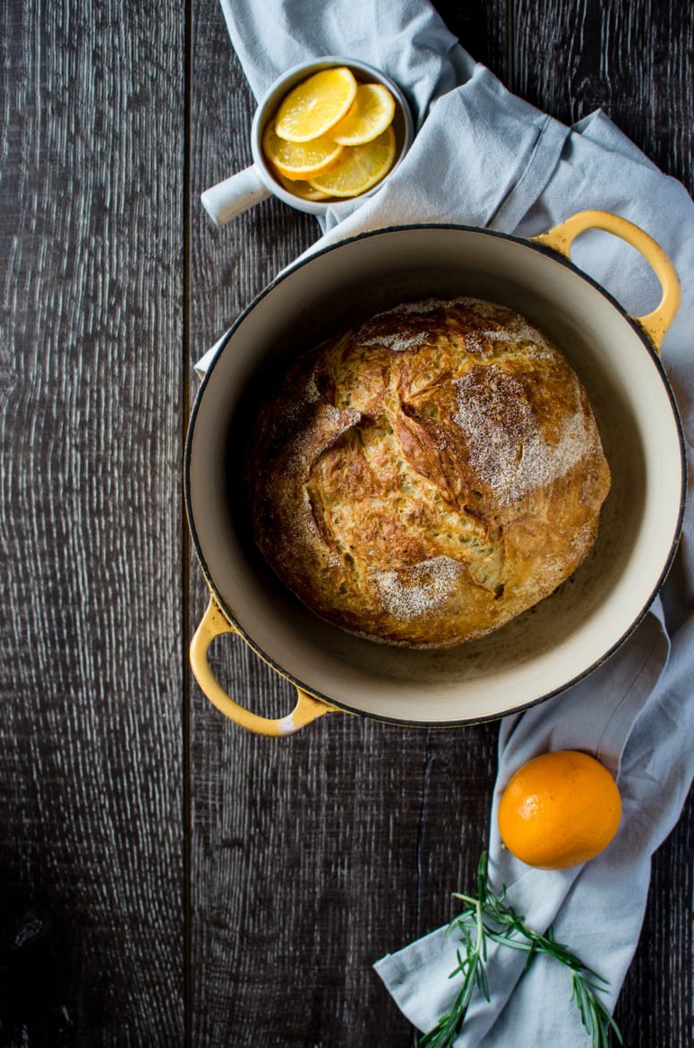 A yellow Dutch oven with a bread boule inside sitting on a grey towel with a bowl of meyer lemons on the side. 