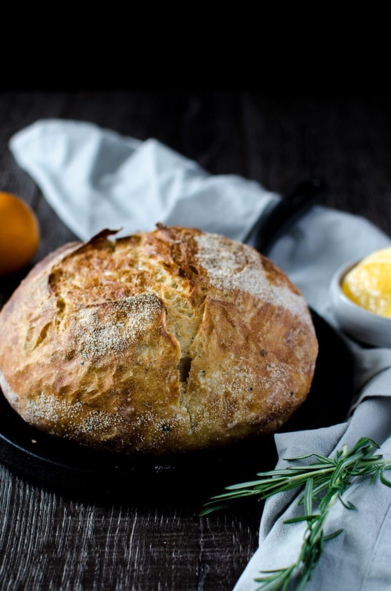 A bread boule sitting on a round cast iron griddle pan with a grey tea towel on the side and a sprig of fresh rosemary.