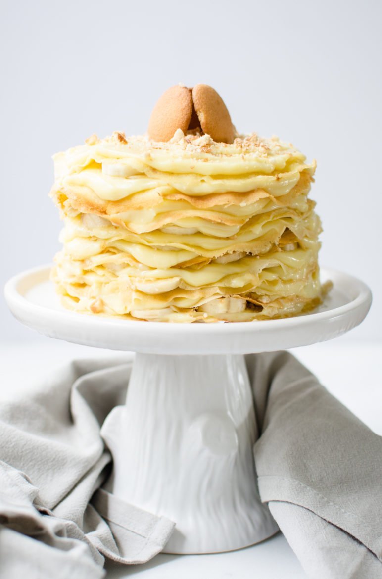 Straight-on shot of a crepe cake with layers of banana pudding, sliced bananas, and Nilla wafers on top of a white ceramic cake stand against a white background. 