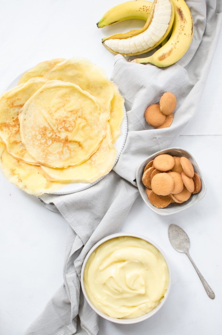 Overhead shot of a plate of crepes, bowl of vanilla pudding, a bunch of bananas, and a bowl of Nilla wafer cookies against a white background. 