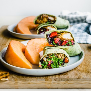 A blue plate with a sliced strawberry pretzel chicken wrap with cantaloupe on the side.