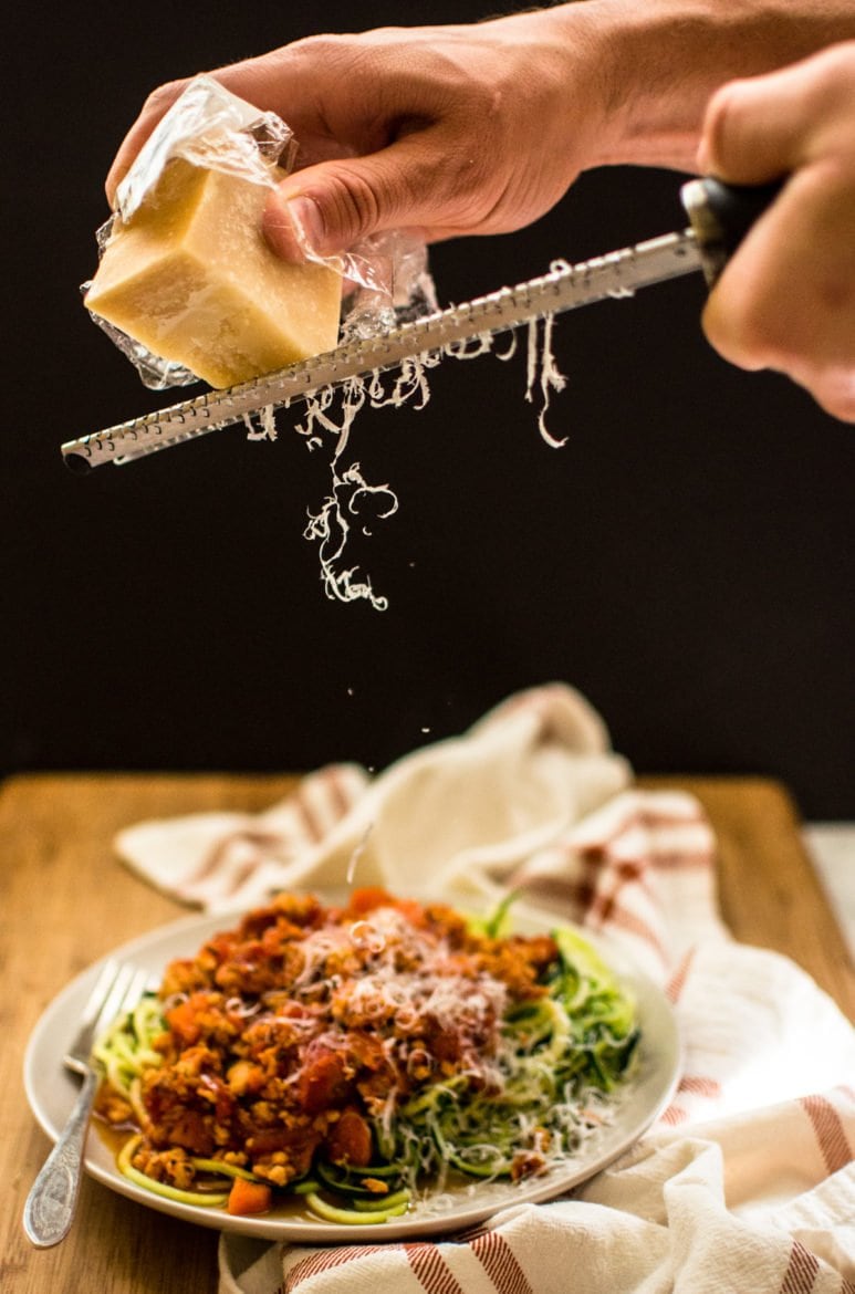 A hand grating Parmesan onto a plate of zucchini noodles topped with bolognese sauce. 