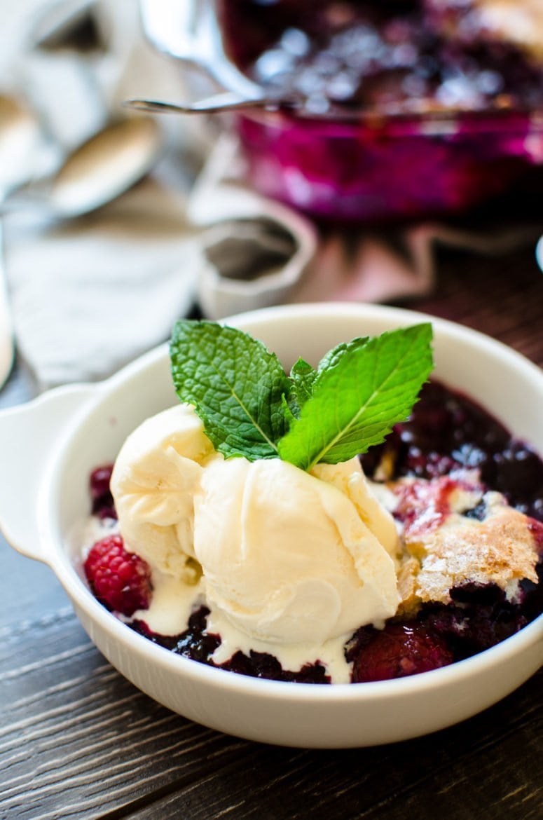A close up shot of a white dish with blueberry cobbler topped with vanilla ice cream and a sprig of fresh mint. 