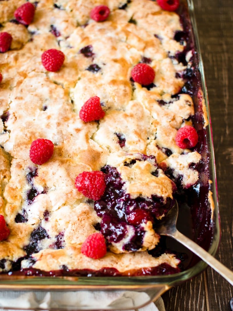 A close up shot of a spoon resting in a pan of baked blueberry cobbler topped with fresh raspberries. 