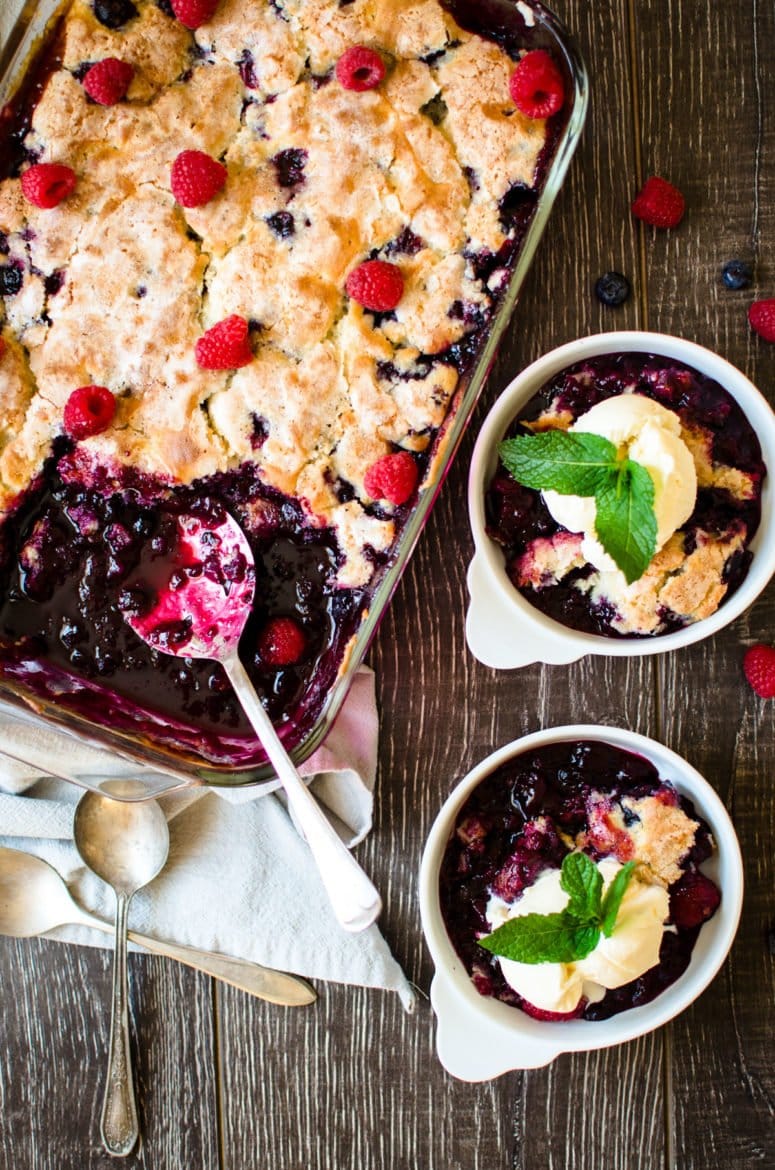 An overhead shot of a pan of blueberry cobbler with two white dishes on the side filled with a serving of cobbler and scoop of ice cream. 