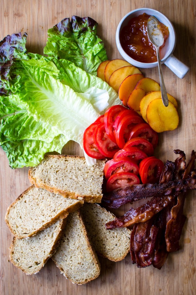 Lettuce, tomatoes, peaches, peach jam, bacon, and bread on a cutting board. 