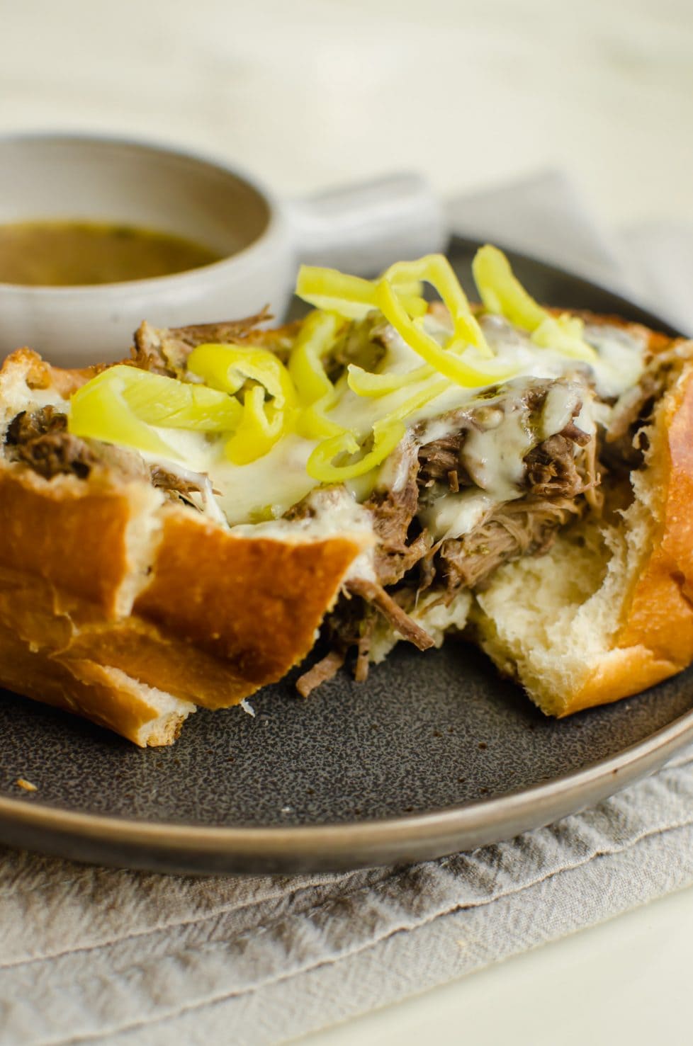 An Italian beef sandwich split in half with strings of white cheese coming out of the center and a cup of au jus on the side. 