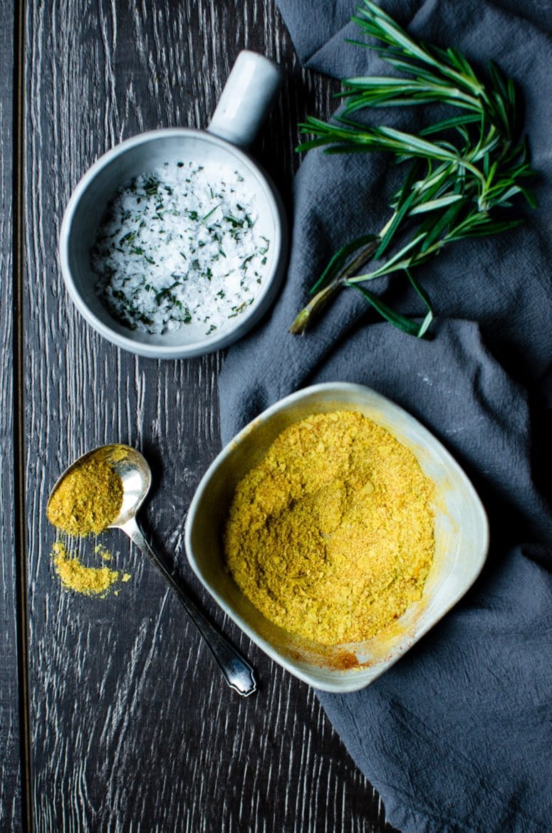 A grey bowl of rosemary sea salt next to a dish of nutritional yeast seasoning. 