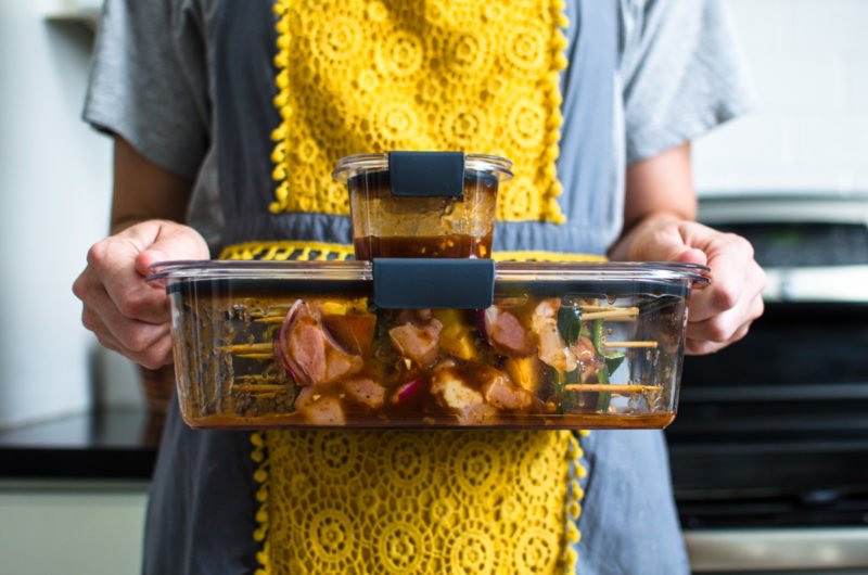 A woman with a grey and yellow apron holding two Rubbermaid Brilliance containers with Peach BBQ chicken kabobs inside. 