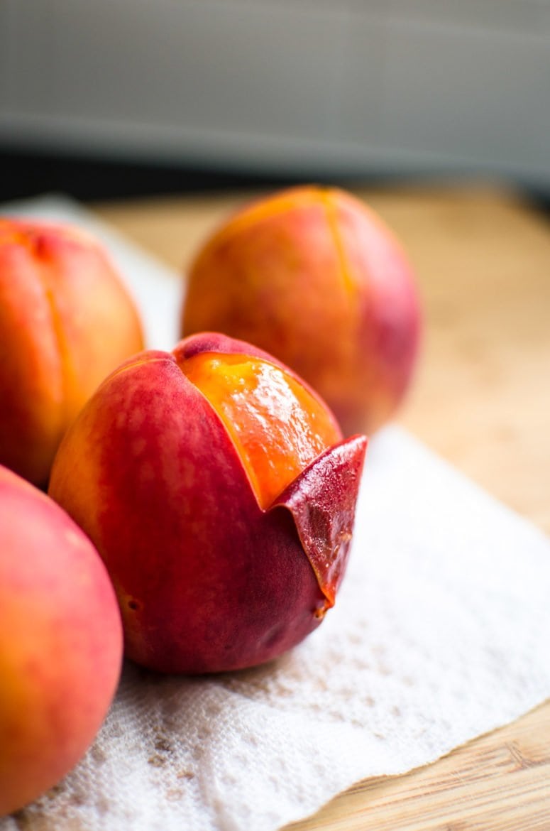 Four poached peaches on a cutting board. One peach has its skin peeled off. 