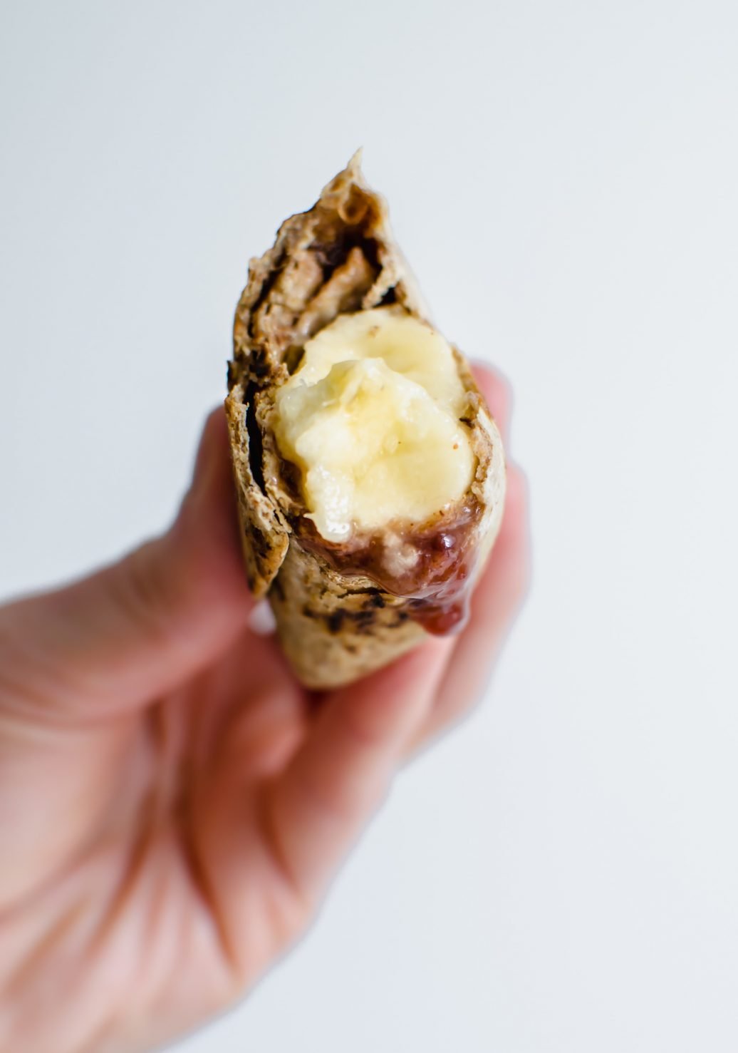 Straight on view of a hand holding half of a whole grain almond butter wrap with a banana inside against a white background.