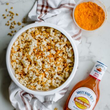 Overhead shot of a white bowl filled with buffalo cheddar popcorn on top of a white marble background with a container of cheddar cheese powder and a bottle of buffalo sauce.