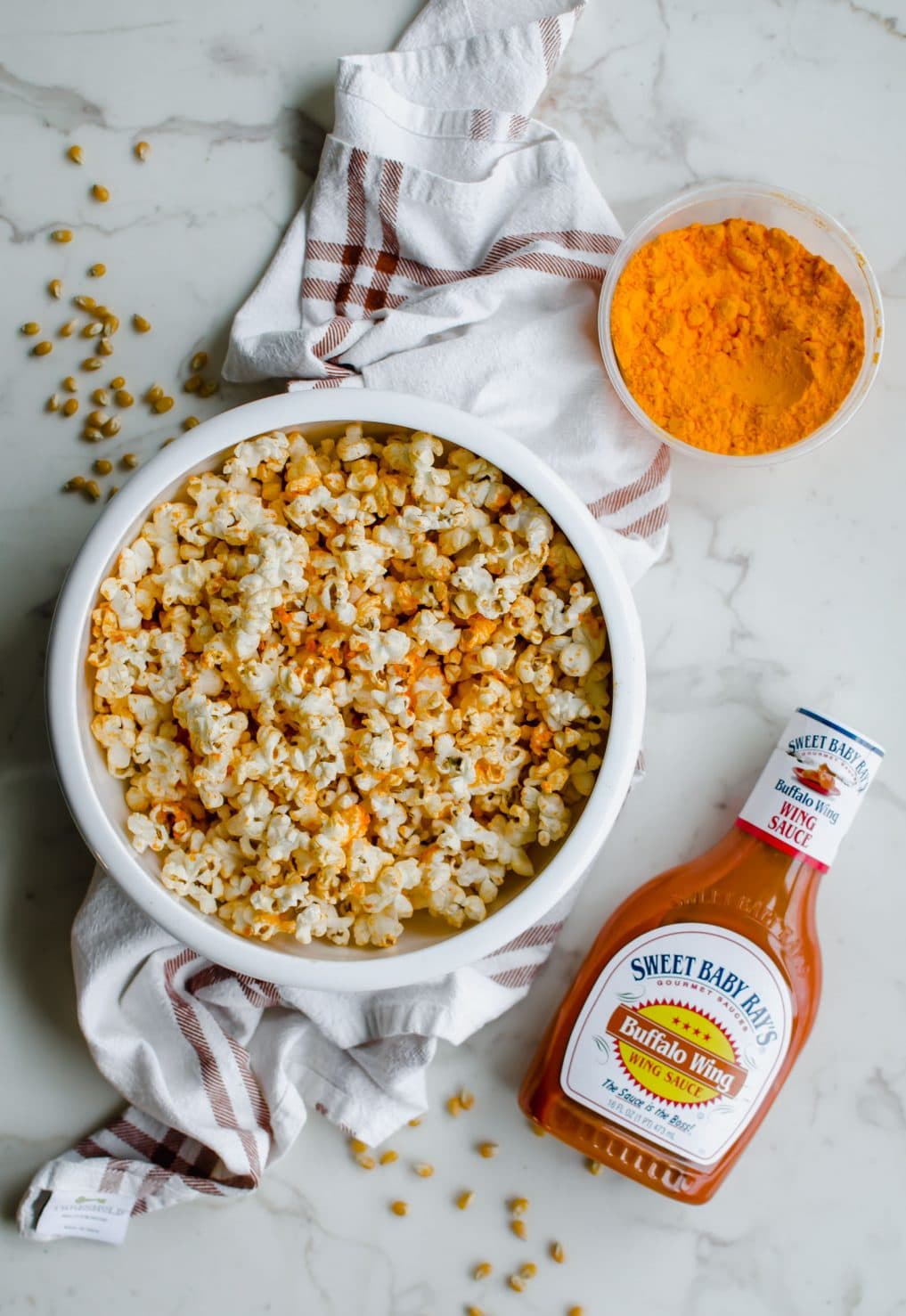 Overhead shot of a white bowl filled with buffalo cheddar popcorn on top of a white marble background with a container of cheddar cheese powder and a bottle of buffalo sauce. 