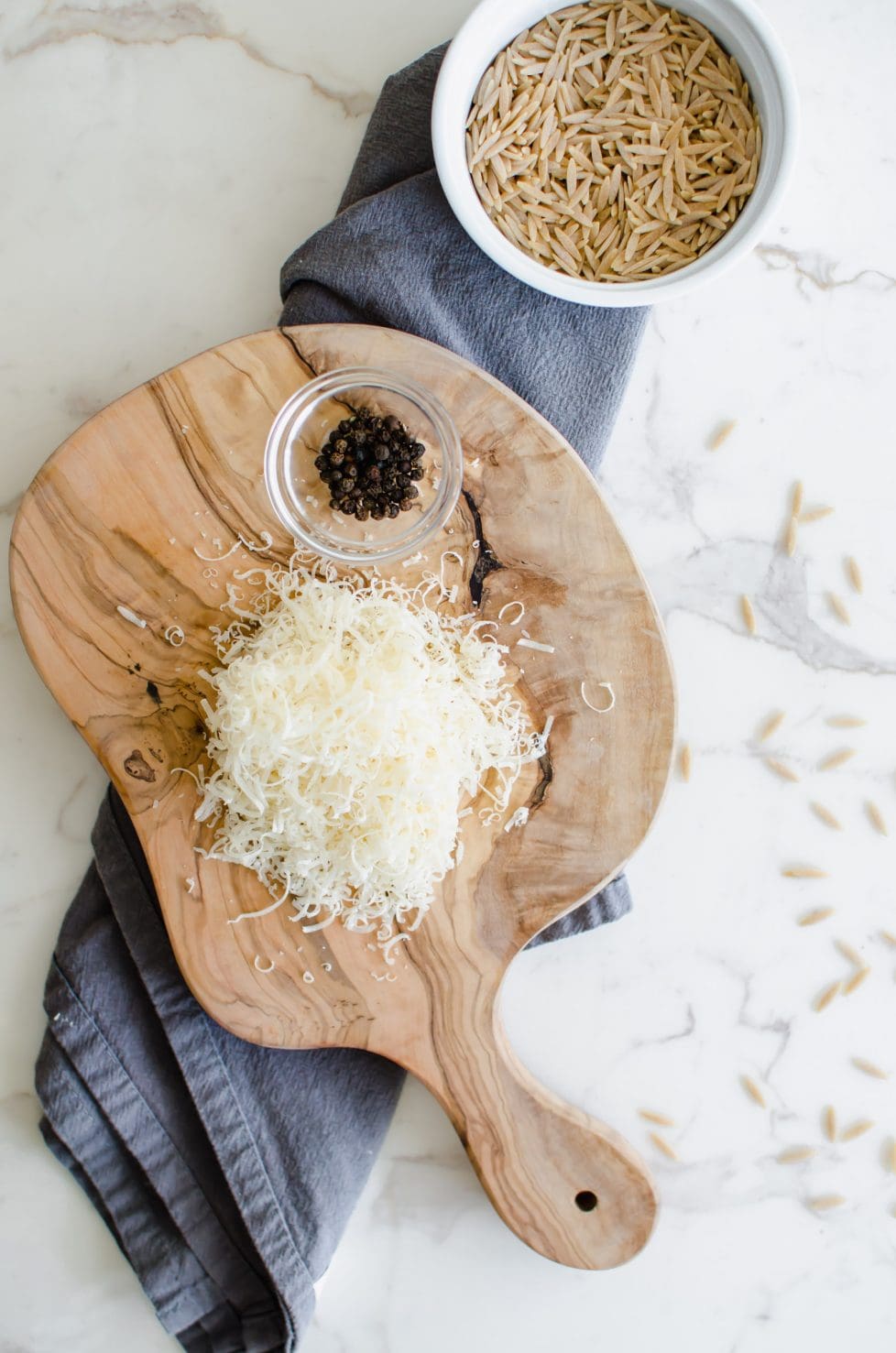 Olive wood cutting board with shredded parmesan cheese and pepper corns sitting on top. Off to the top there is a bowl of uncooked orzo pasta.