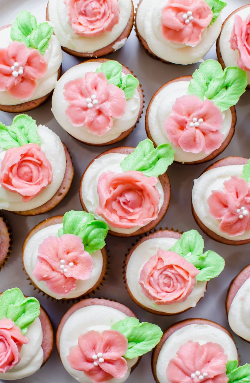 An overhead shot of many strawberry cupcakes with cream cheese frosting that are topped with pink and green buttercream flowers. 