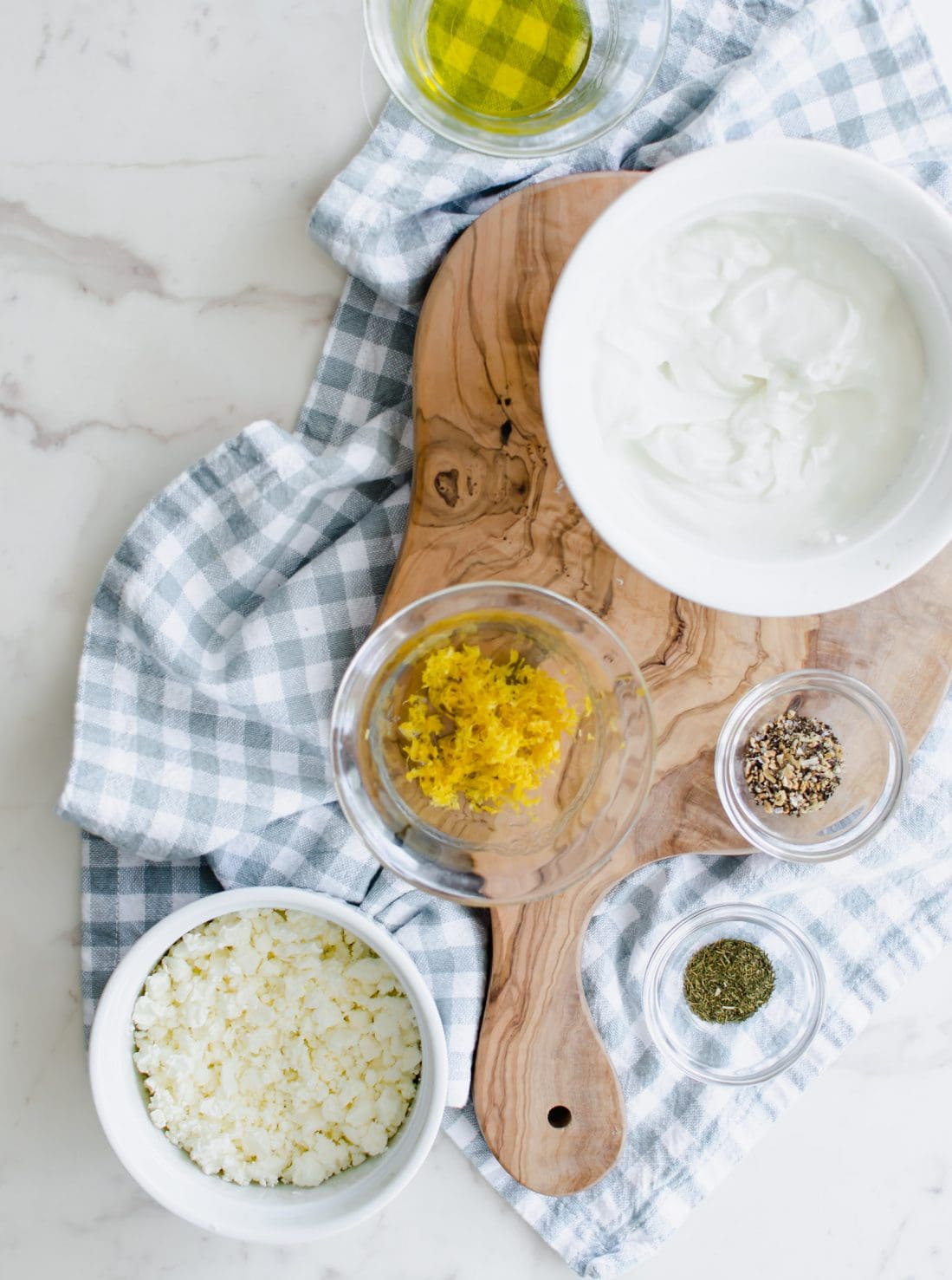 An overhead shot of bowls with ingredients for making a feta dipping sauce on a white marble counter top. 