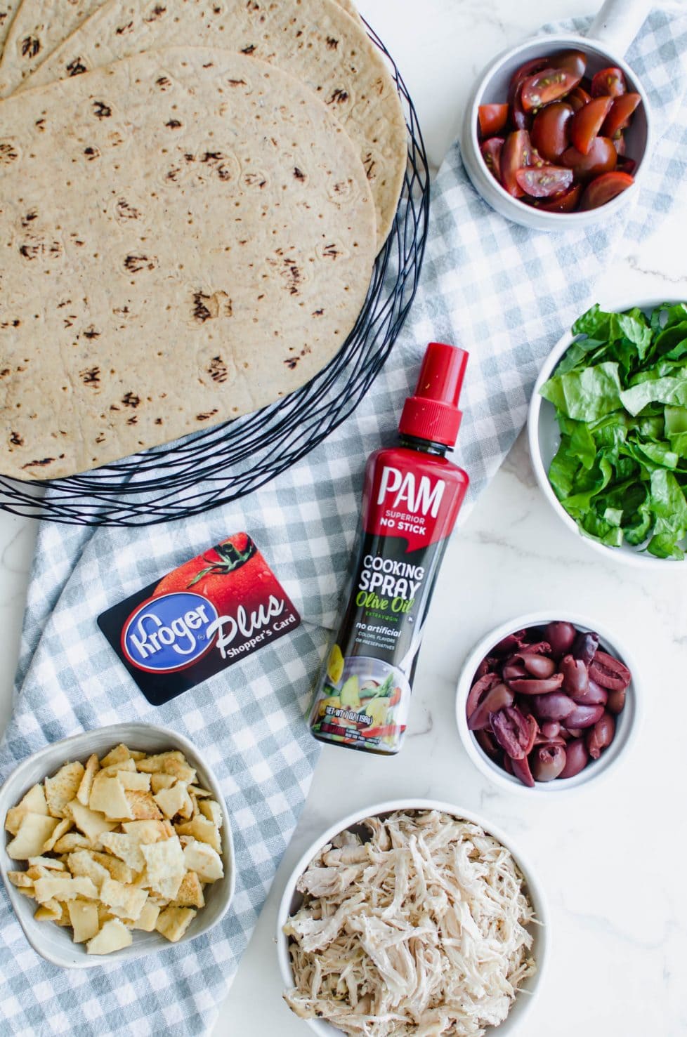 An overhead shot of bowls of tomatoes, lettuce, shredded chicken, pita chips, olives, a plate of whole grain wraps, and a bottle of Pam olive oil cooking spray against a white background. 