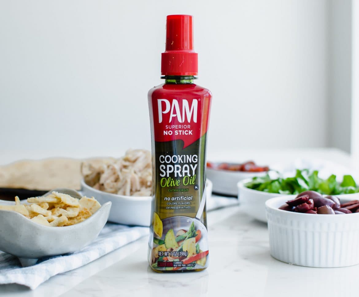 A bottle of PAM non-aerosol olive oil spray with white bowls of Greek salad toppings on a white marble counter. 