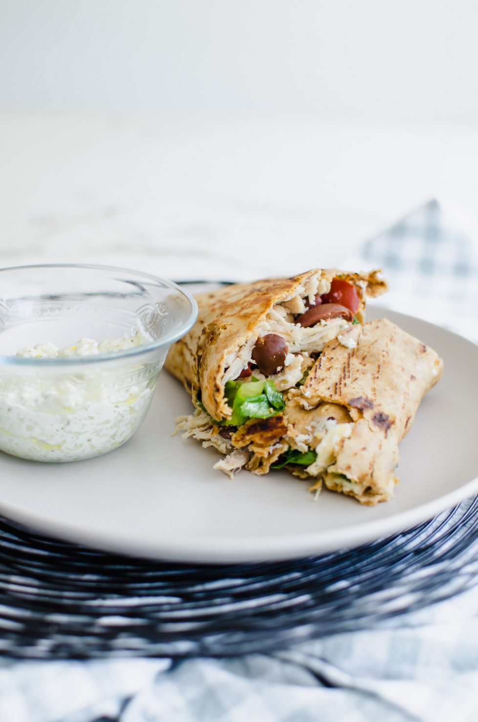 A close-up shot of a Greek Chicken Crunchwrap sliced in half on a white plate with a glass bowl of feta dipping sauce.