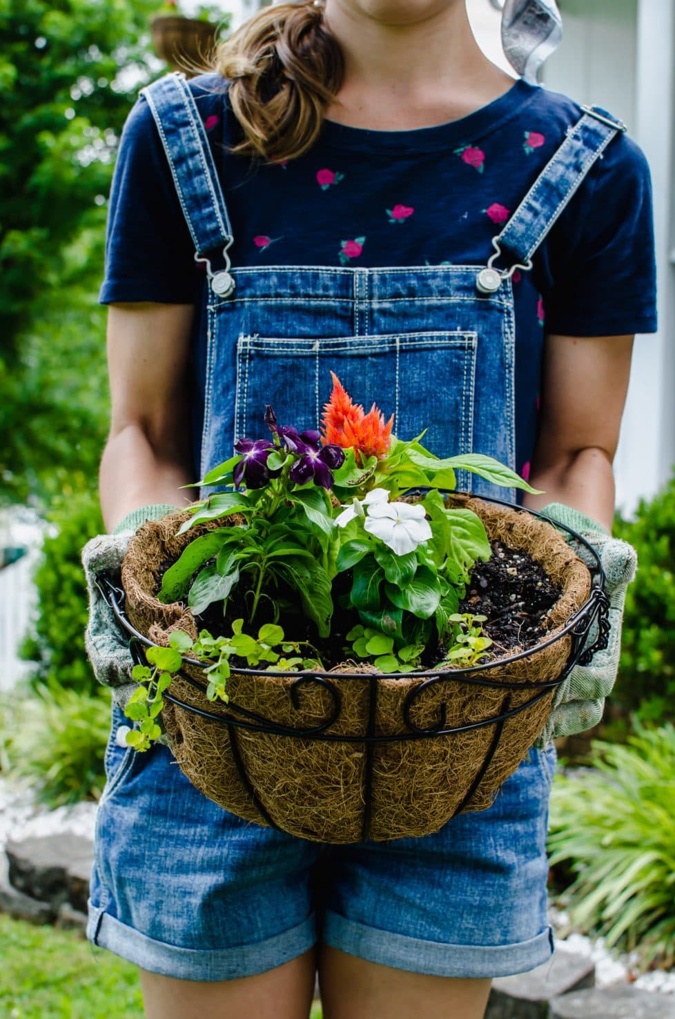 A straight-on shot of a girl holding a hanging flower basket filled with coral, purple, and white flowers. 