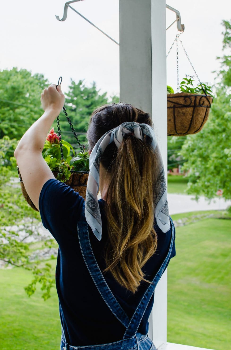 A shot of the back of a girl hanging a flower basket on a front-porch hook.