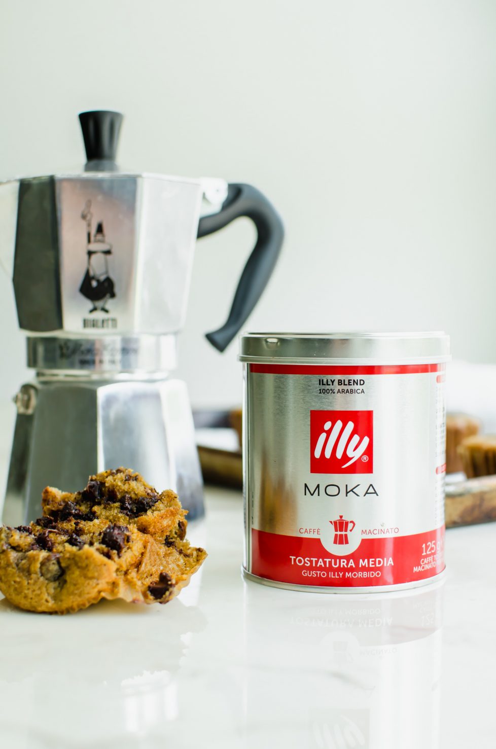 A straight-on shot of a can of Illy espresso, a stovetop coffee maker, and a chocolate chip muffin broken in half.