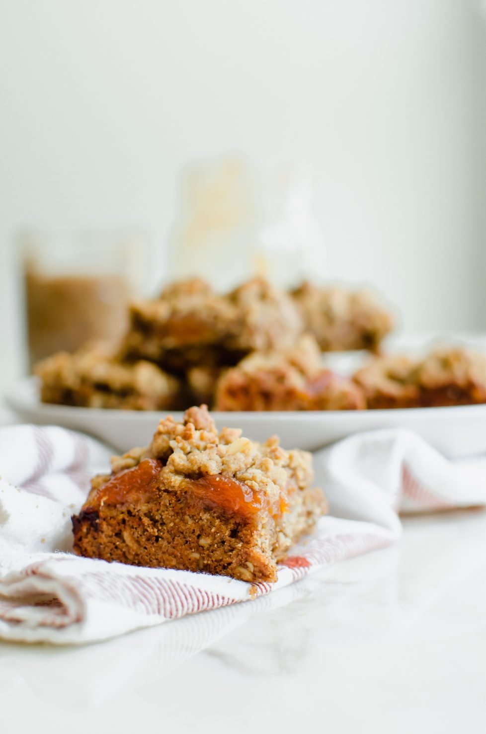 A side shot of a Pecan Butter and Peach Jam Crumble Bar sitting on a plaid kitchen towel with a white plate of bars in the background. 