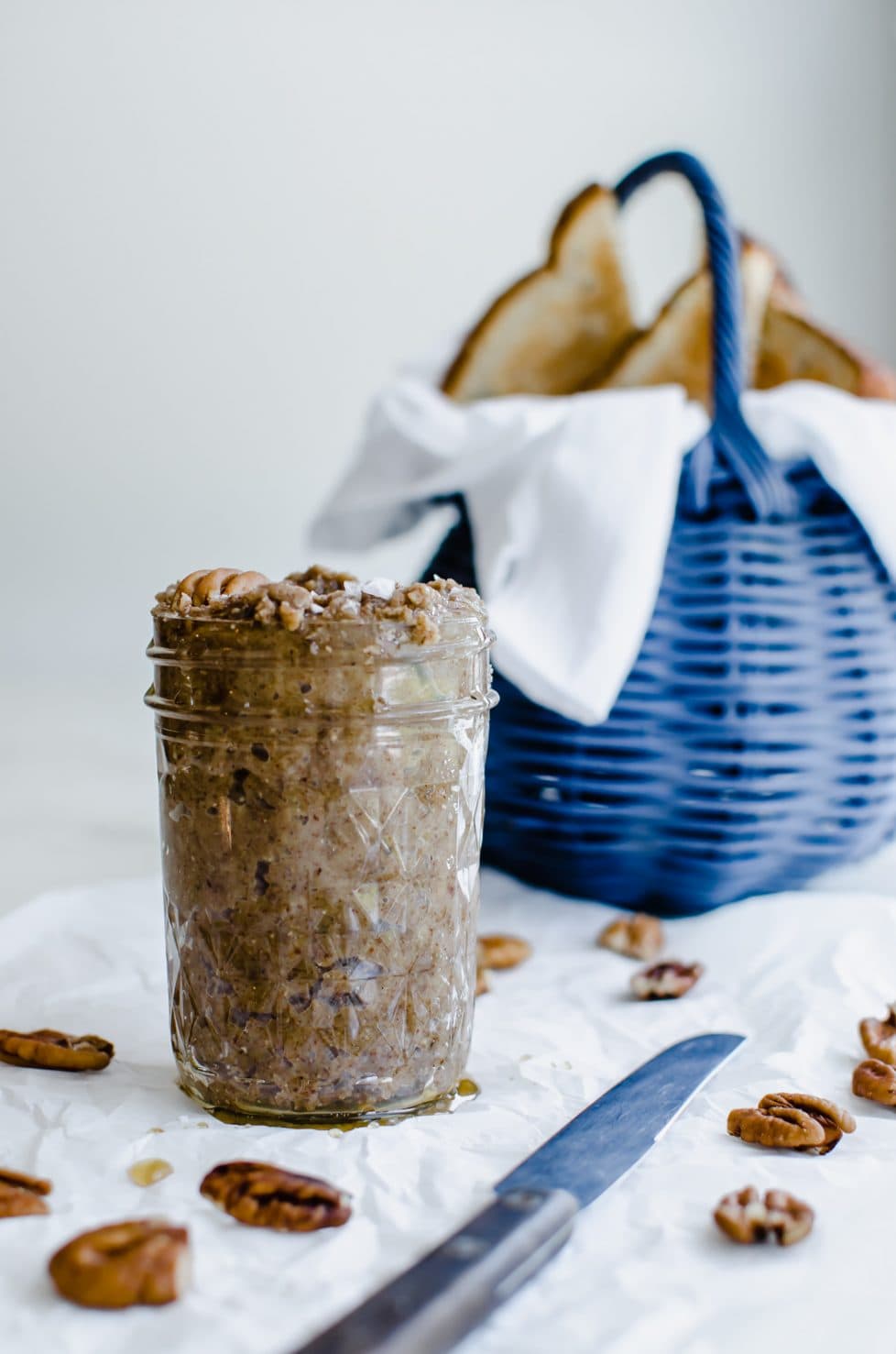 A side shot of a jar of salted maple pecan butter with a blue basket in the background that is filled with toast.