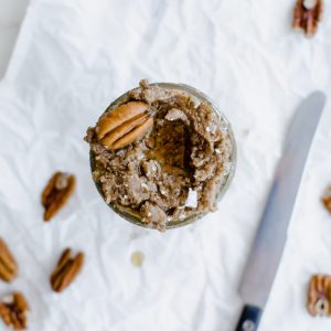 An overhead shot of a mason jar with maple pecan butter inside. The top is garnished with a pecan halve and some sea salt flakes.