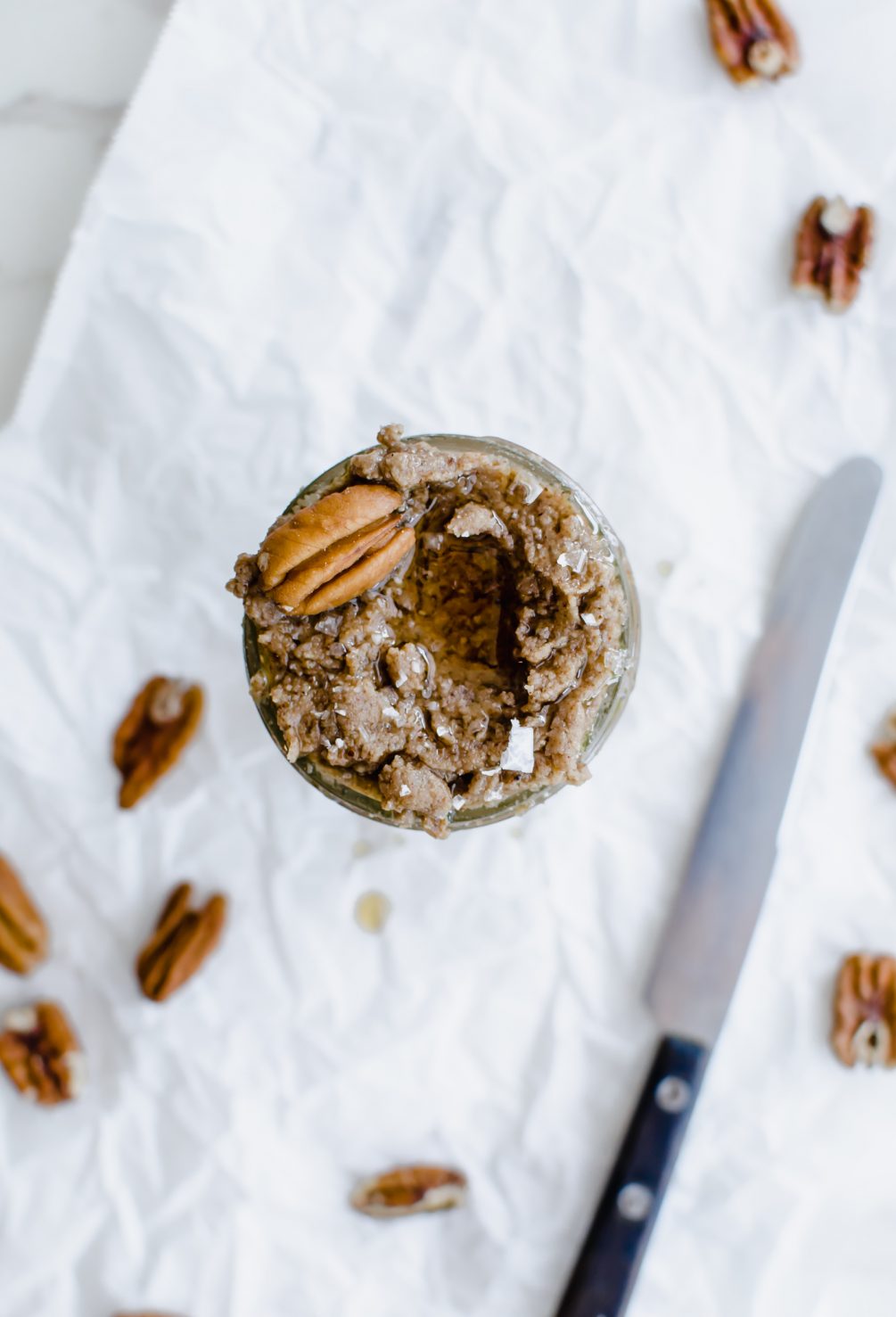 An overhead shot of a mason jar with maple pecan butter inside. The top is garnished with a pecan halve and some sea salt flakes.