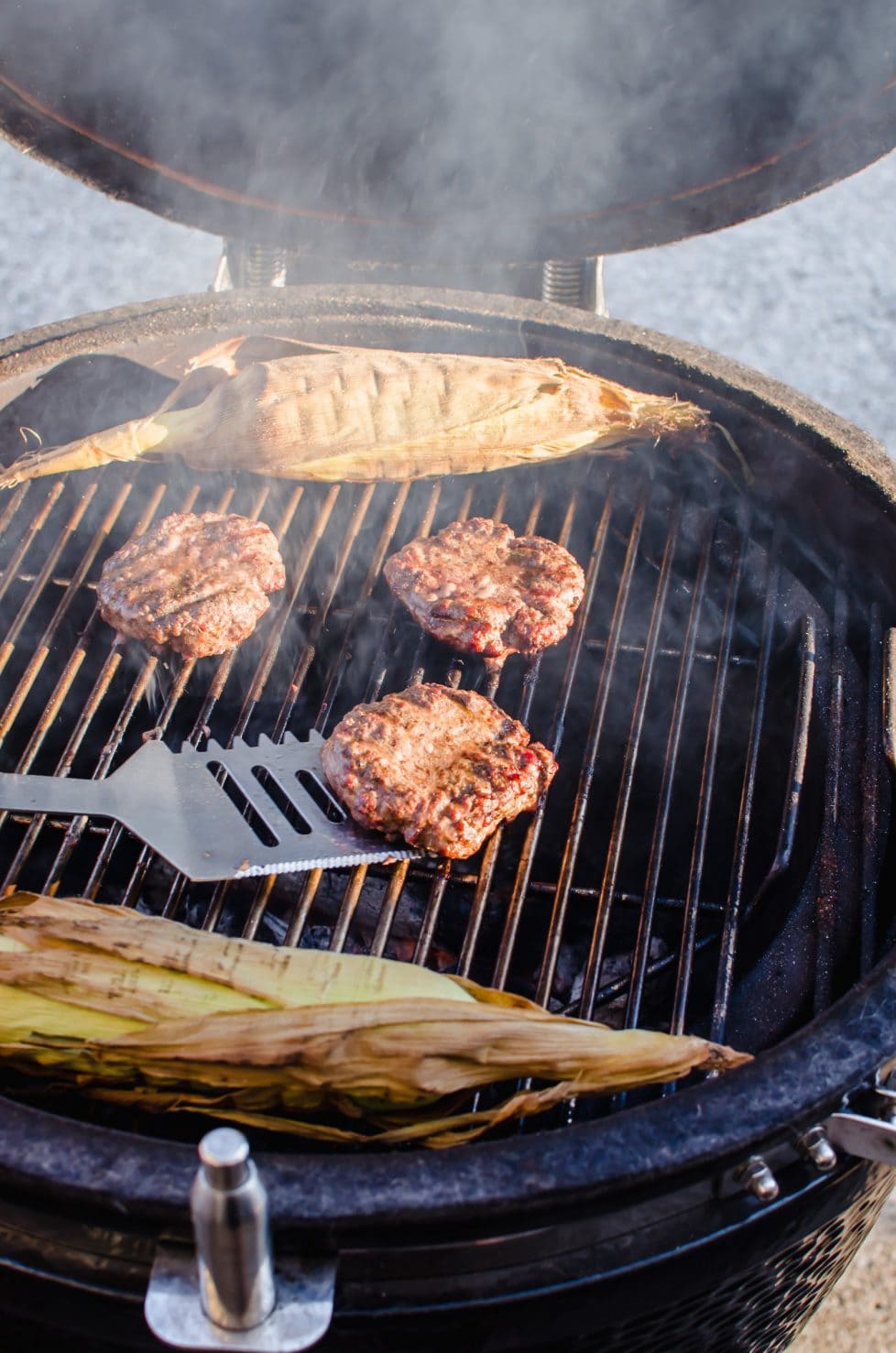 Burger patties being flipped on a grill with two ears of corn in the shuck.