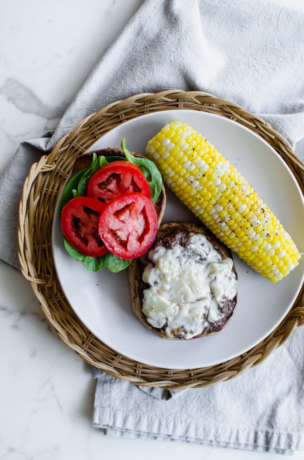 A view from above of an open-faced burger with sliced tomato, spinach, white cheddar, and an ear of corn on the side. 