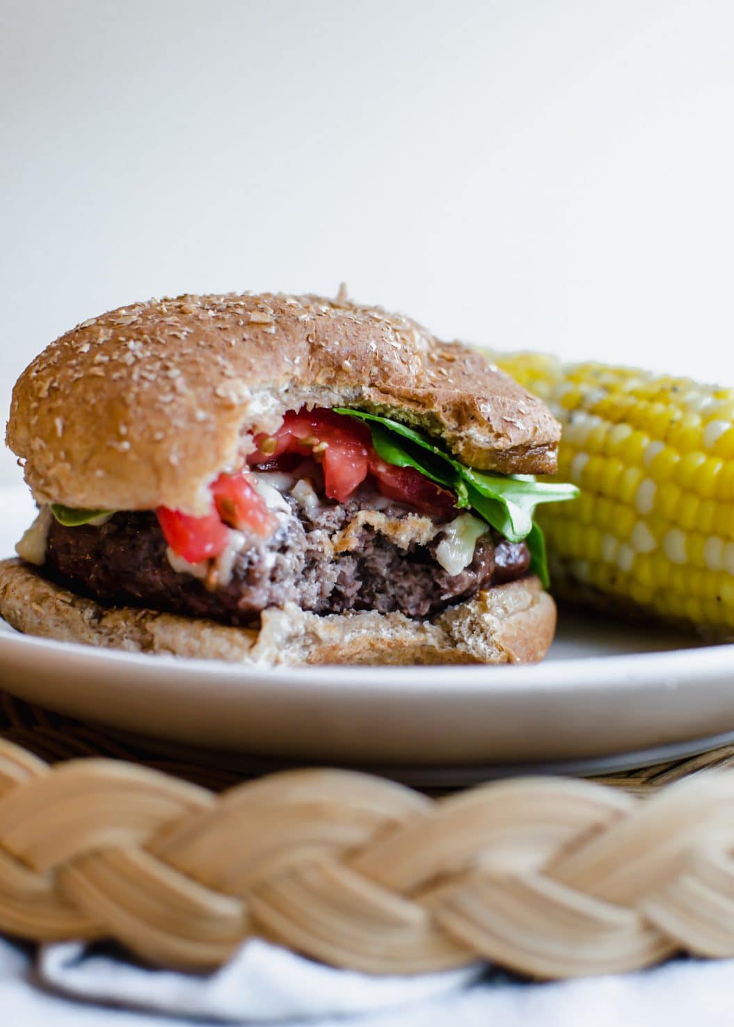 A burger topped with spinach and tomato with a bite taken out of it and an ear of corn on the side. 